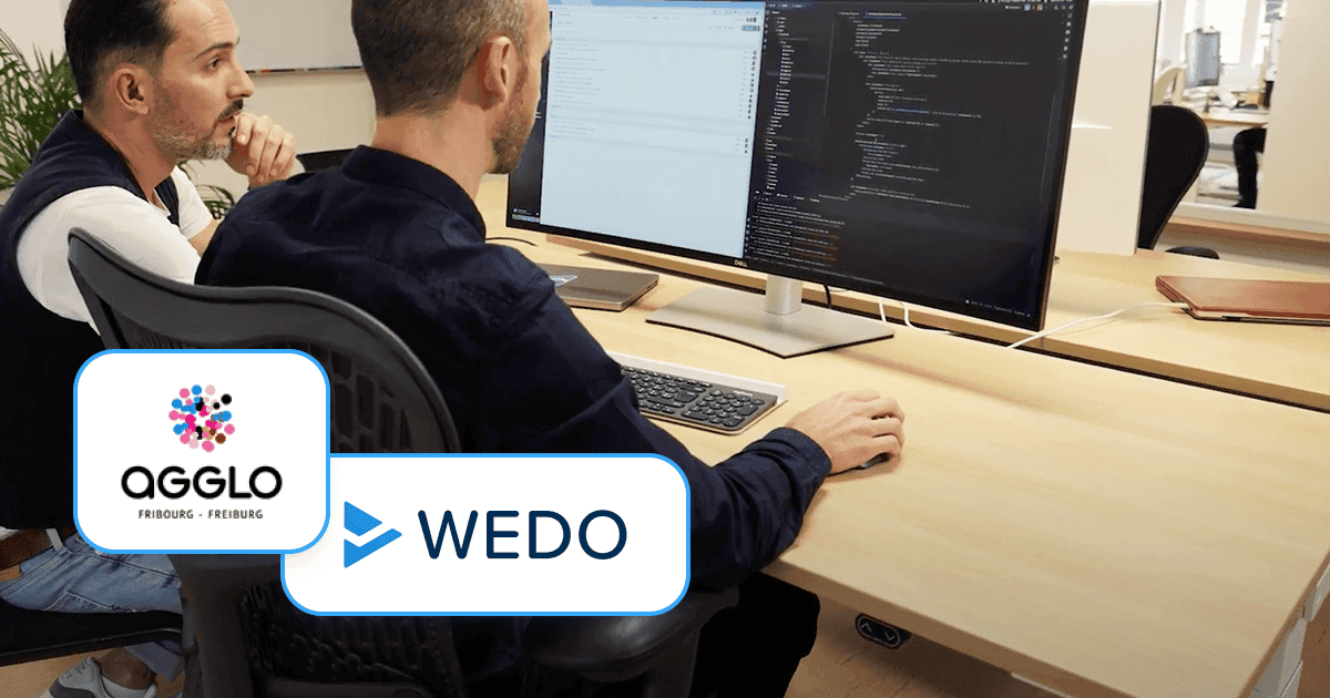 Two colleagues sit side by side at a desk, viewing lines of code and a project interface displayed on a large computer monitor. In the bottom left corner, the AGGLO Fribourg–Freiburg logo and the WEDO logo are overlaid.